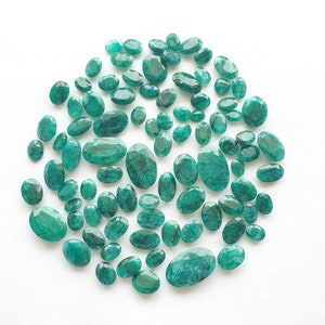 May include: A collection of emerald green gemstones of various sizes and shapes, arranged in a circular pattern on a white background.