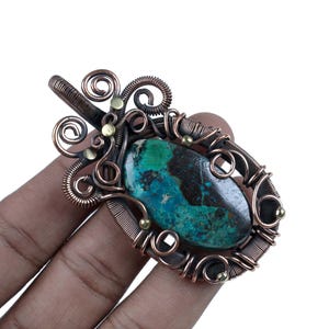 Puede incluir: Colgante artesanal con una piedra preciosa ovalada pulida con tonos turquesa y marrón, engastada en un marco de alambre de cobre. El trabajo de alambre incluye espirales decorativas y pequeños detalles de latón, creando una pieza de joyería artística única.