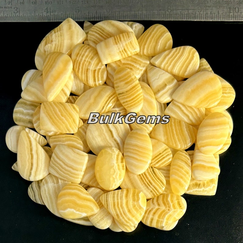 May include: A collection of yellow and white banded agate cabochons in various shapes, including teardrop, oval, and rectangular. The stones are arranged in a pile on a black background. BulkGems is printed on the image.
