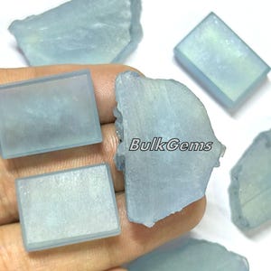 Può includere: Una collezione di sei grezzi di cristallo di acquamarina blu chiaro, alcuni sono rettangolari e altri hanno forme irregolari. I cristalli sono traslucidi e hanno un leggero bagliore iridescente. BulkGems è stampato sull'immagine.