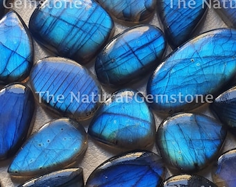 May include: A collection of blue labradorite gemstone cabochons, each with a unique iridescent sheen. The stones are oval and teardrop shaped, and they are arranged in a random pattern. The Natural Gemstone is printed on some of the stones.