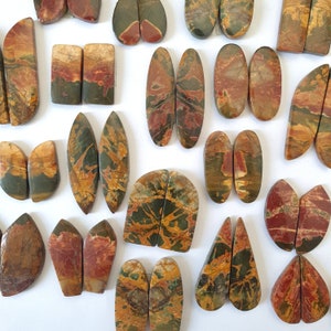 Puede incluir: Una colección de cabujones de piedra natural pulida en varias formas, incluyendo óvalos, rectángulos y lágrimas. Las piedras muestran una mezcla de colores terrosos: marrones, verdes y rojos, con patrones de vetas únicos. Ideal para joyería o artesanía.