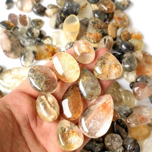 May include: A collection of polished, translucent gemstones in various shapes and sizes, including oval and teardrop forms. The stones display a range of colors, from clear to golden brown, with internal inclusions. Some stones are held in a hand, while others are scattered.