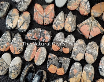 May include: A collection of natural gemstone cabochons in various shapes and sizes. The cabochons are a mix of black, white, gray, and red with gold flecks. The cabochons are arranged on a black surface. The Natural Gemstone is printed on the image.