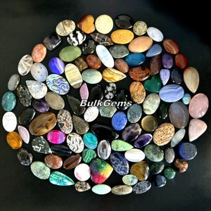 May include: A collection of approximately 100 assorted gemstone cabochons in various shapes, sizes, and colours. The cabochons are arranged in a circular pattern on a black background. The text "BulkGems" is visible in the centre of the image.