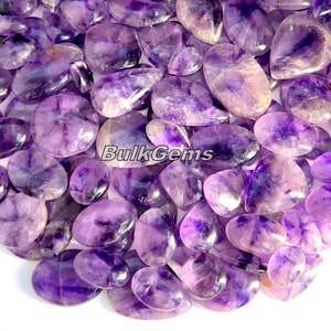 May include: A close-up view of a pile of purple amethyst gemstone cabochons. The cabochons are smooth and polished, with a variety of shapes and sizes. The stones are a deep purple color, with some having a lighter or darker hue. The cabochons are arranged in a random pattern, with some overlapping.