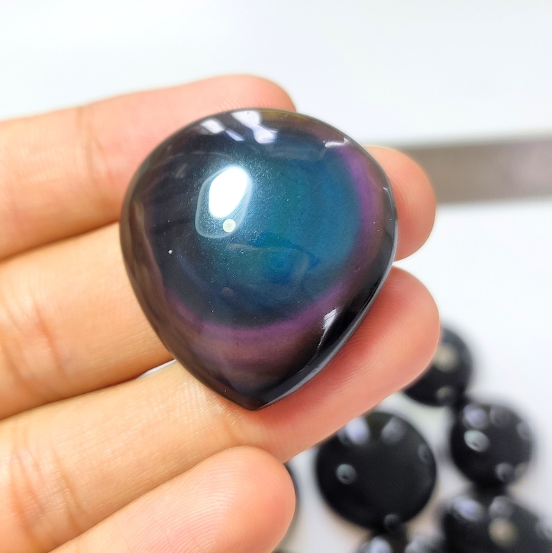 K&ouml;nnte beinhalten: Ein dunkler, regenbogenfarbener herzf&ouml;rmiger Stein mit einem blauen, gr&uuml;nen und violetten Schimmer. Der Stein wird in einer Hand gehalten.