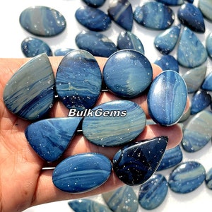 May include: A collection of blue and grey polished gemstone cabochons with white flecks. The cabochons are oval, teardrop, and triangular shaped. The cabochons are arranged on a hand.