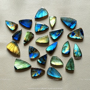 Op de afbeelding: Een verzameling van 20 driehoekige labradorit-cabochons in tinten blauw, groen en goud. De stenen zijn in een cirkelvormige opstelling op een lichtbruin oppervlak geplaatst.