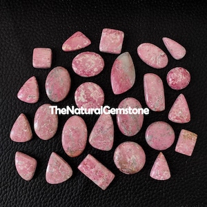 May include: A collection of 20 pink and white gemstone cabochons in various shapes and sizes. The gemstones are arranged on a black surface. TheNaturalGemstone is printed on the surface.