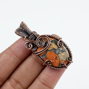 Puede incluir: Colgante ovalado con alambre de cobre. El colgante presenta una piedra con una mezcla de colores naranja, gris y marrón. El trabajo de alambre incluye espirales decorativas y una sección superior tejida, creando una pieza de joyería única.