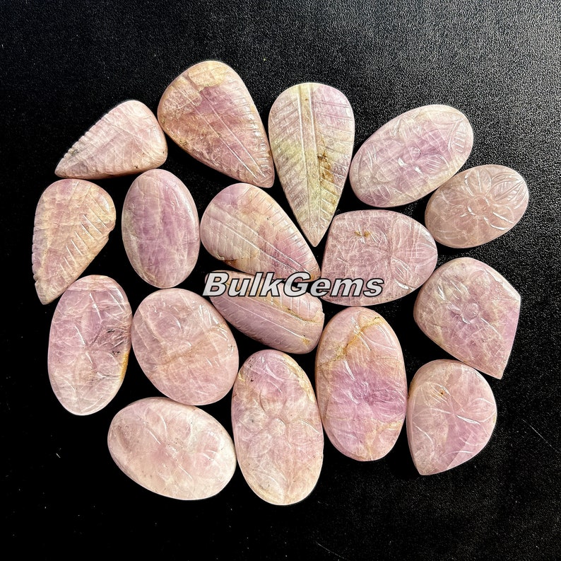 May include: A collection of 15 pink and white carved gemstone cabochons. The cabochons are oval and teardrop shaped and have a variety of carved designs. The cabochons are arranged in a circle on a black background.