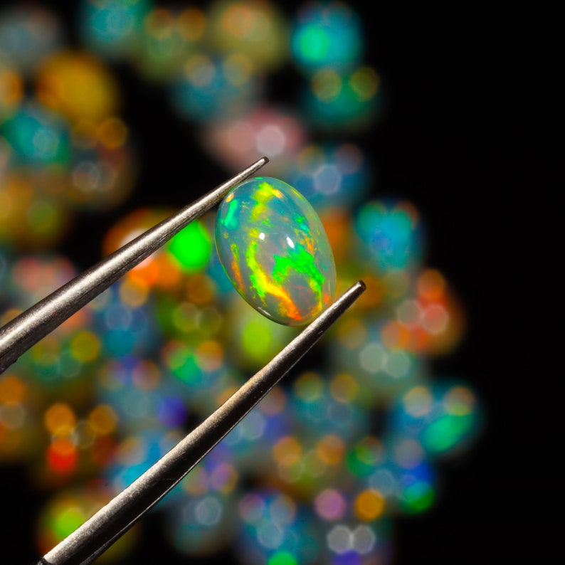 May include: A green and blue oval opal gemstone held by tweezers against a background of out-of-focus opals.