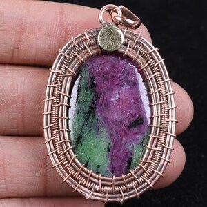 Peut inclure: Un pendentif ovale composé d'une pierre précieuse de rubis zoïsite polie avec des inclusions magenta et vertes vibrantes. La pierre est enchâssée dans un cadre en fil de cuivre travaillé avec un accent métallique circulaire et une bélière en cuivre.
