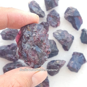 May include: A close-up of a hand holding a dark red, rough, and irregularly shaped gemstone. The gemstone has a slightly translucent quality and appears to be a raw, unpolished piece of garnet. The background shows several other similar gemstones scattered on a white surface. The text "BulkNaturalGemstone" is visible in the lower right corner of the image.