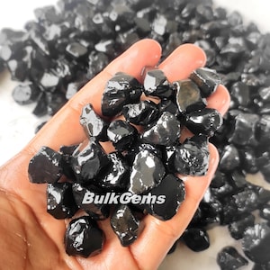 May include: A hand holding a pile of black gemstones. The gemstones are rough and have a shiny, polished finish. The text "BulkGems" is visible in the image.