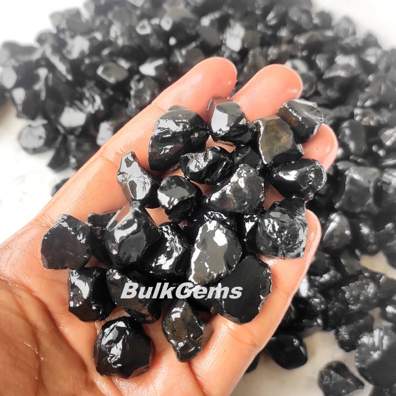May include: A hand holding a pile of black gemstones. The gemstones are rough and have a shiny, polished finish. The text "BulkGems" is visible in the image.