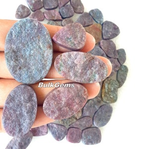May include: A collection of oval and triangular shaped cabochons of blue and pink ruby. The stones have a mottled, cloudy appearance. The stones are arranged on a white surface. BulkGems is printed on the image.