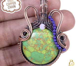 May include: A handcrafted copper wire pendant with a round, green gemstone. The gemstone has brown and yellow veining. The pendant is adorned with purple beads and a small white bead. The image includes the text "Bulk Gems".