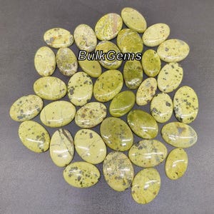 May include: A collection of green and brown oval-shaped stones with a mottled pattern. The stones are arranged in a random pattern on a gray surface. The text "BulkGems" is visible in the center of the image.