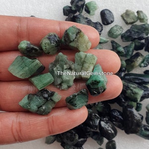 May include: A hand holding a collection of rough, green emerald gemstones. The stones are various sizes and shapes, with some having black inclusions. TheNaturalGemstone is printed on the image.