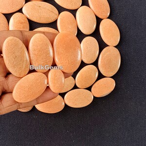May include: A collection of oval-shaped, peach-colored gemstones. Some are held in a hand, while others are scattered on a dark surface. The gemstones have a smooth, polished appearance and a subtle, natural grain.