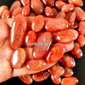 Puede incluir: Una colección de cabujones de piedra solar de forma ovalada, pulidos, en tonos naranja y rojo. Las piedras tienen un acabado brillante e iridiscente.