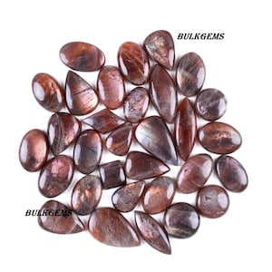 Op de afbeelding: Een verzameling gepolijste zonnesteen edelstenen in verschillende vormen, waaronder ovaal, druppelvormig en marquise. De stenen vertonen een mix van roodbruine en iriserende kleuren, met een sprankelend effect. De tekst "BULKGEMS" is zichtbaar.