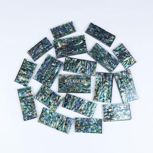 Może przedstawiać: Kolekcja 17 iryzujących płytek z muszli abalone w różnych odcieniach niebieskiego, zielonego i fioletowego. Płytki są prostokątne i mają naturalną, organiczną fakturę. Płytki są ułożone w okrąg na białym tle. Słowo "BULKGEEMS" jest nadrukowane na biało na jednej z płytek.