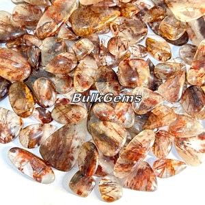 May include: A collection of raw, natural, red and gold, rutilated quartz gemstones. The stones are in various shapes and sizes, with a clear, translucent appearance. The rutile inclusions create a shimmering effect within the quartz.