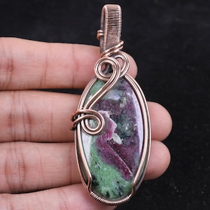 Peut inclure: Un pendentif de forme ovale composé d'une pierre précieuse de rubis zoïsite polie avec des inclusions magenta profond et vert forêt. La pierre est entourée d'un fil de cuivre décoratif avec des spirales et une bélière supérieure.