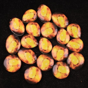 Pode incluir: Uma coleção de vinte e um cabochões com um design celestial. Cada cabochão apresenta um núcleo central amarelo rodeado por uma mistura de tons laranja e castanho, assemelhando-se a uma nebulosa ou galáxia. Os cabochões variam em forma, incluindo oval e redondo.