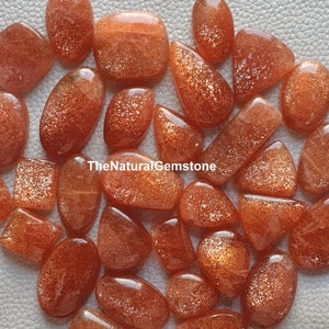 May include: A collection of 20+ sunstone cabochons in various shapes and sizes. The stones are a mix of orange and red with a shimmery, iridescent finish. TheNaturalGemstone is printed on the image.