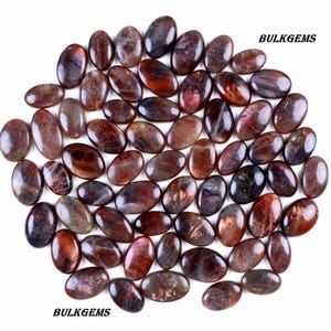 May include: A collection of oval-shaped sunstone gemstones in various shades of brown, red, and black, with a shimmering, iridescent effect. The gemstones are arranged closely together on a white background. The text "BULKGEMS" is visible.