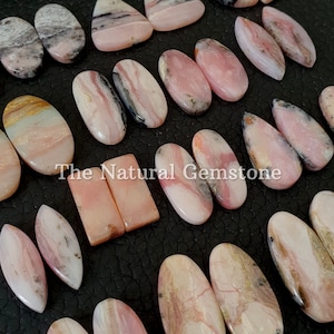 Puede incluir: Una colección de cabujones de ópalo rosa y blanco con marcas negras y marrones. Los cabujones son de varias formas y tamaños, incluyendo ovalados, en forma de lágrima y rectangulares. El texto "The Natural Gemstone" está impreso en la imagen.