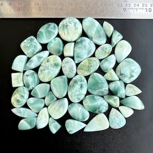 May include: A collection of 30 green and blue gemstone cabochons, with a variety of shapes and sizes. The cabochons are arranged in a semi-circle on a black surface.