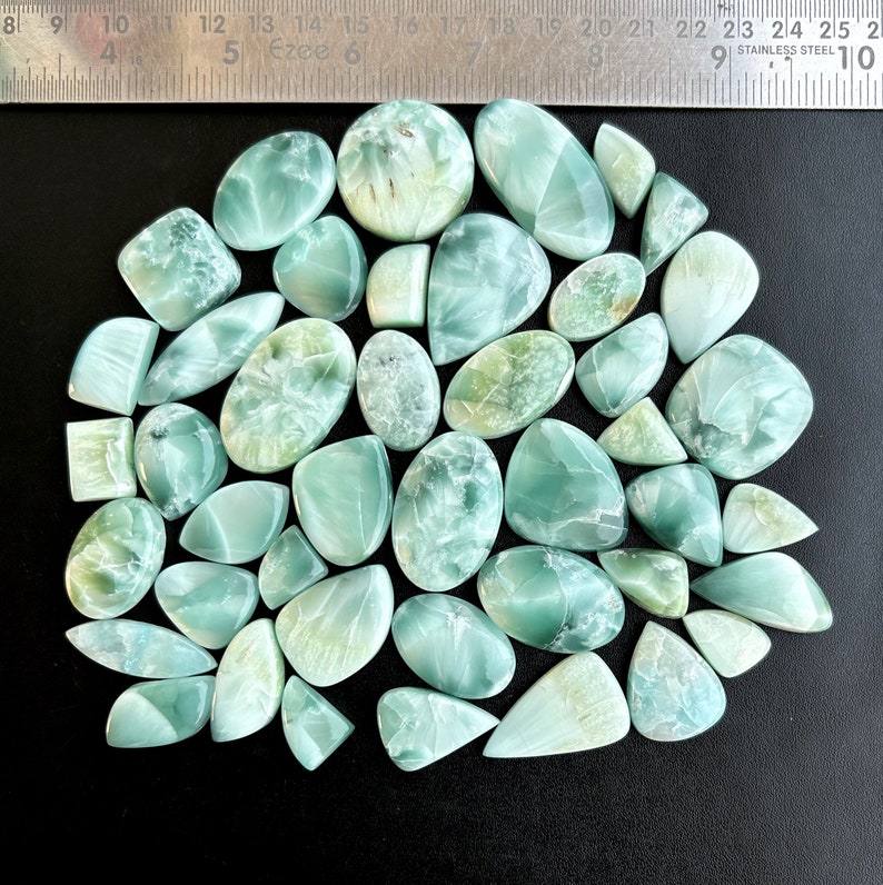May include: A collection of 30 green and blue gemstone cabochons, with a variety of shapes and sizes. The cabochons are arranged in a semi-circle on a black surface.