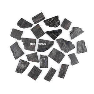Puede incluir: Colección de piezas de turmalina de color gris oscuro a negro, de forma irregular, dispersas sobre un fondo blanco. Las piezas varían en tamaño y textura, con estrías visibles. El texto "BULKGEMS" es visible.