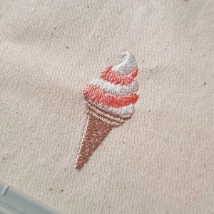 May include: Embroidered ice cream cone design on a beige fabric. The ice cream is pink and white with a brown cone.
