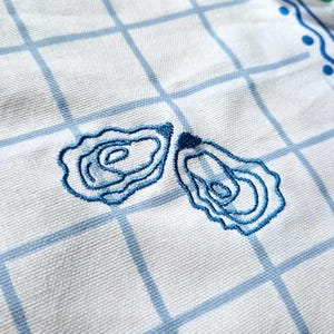 Puede incluir: Pa&ntilde;o de cocina blanco con un patr&oacute;n de cuadr&iacute;cula azul y bordados de conchas de ostra azules. La toalla tiene un borde decorativo con puntos azules y hojas verdes. La tela es tejida.