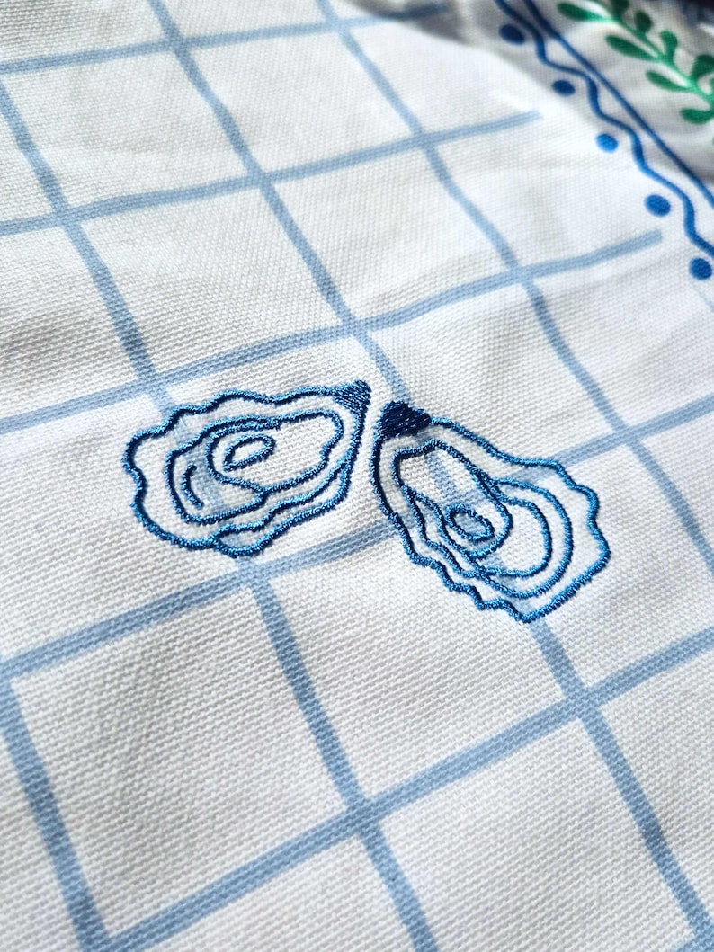 Puede incluir: Pa&ntilde;o de cocina blanco con un patr&oacute;n de cuadr&iacute;cula azul y bordados de conchas de ostra azules. La toalla tiene un borde decorativo con puntos azules y hojas verdes. La tela es tejida.