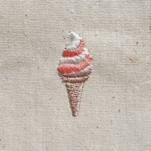 May include: Embroidered ice cream cone with pink and white swirls on a beige fabric background.