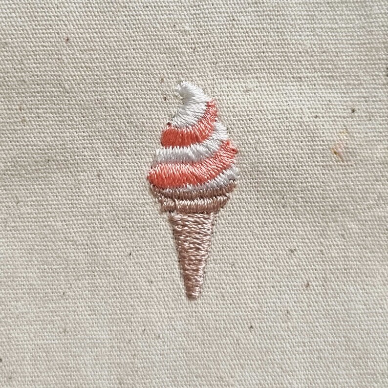 May include: Embroidered ice cream cone with pink and white swirls on a beige fabric background.