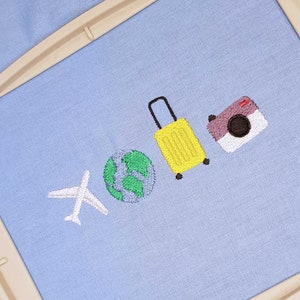 May include: Embroidered travel-themed design on light blue fabric. Features a white airplane, a green and blue globe, a yellow suitcase, and a purple camera. The design is likely for a patch or embroidery project.