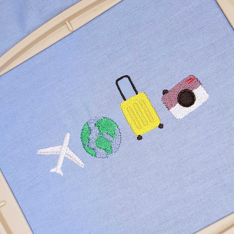 May include: Embroidered travel-themed design on light blue fabric. Features a white airplane, a green and blue globe, a yellow suitcase, and a purple camera. The design is likely for a patch or embroidery project.