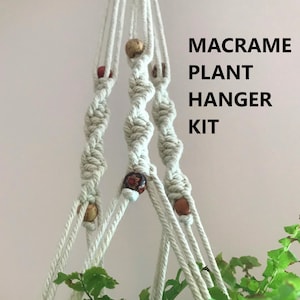 May include: A macrame plant hanger kit featuring off-white cotton cord knotted in a spiral pattern with decorative wooden beads in various brown and red tones. The text MACRAME PLANT HANGER KIT is printed in black on the right side of the image.