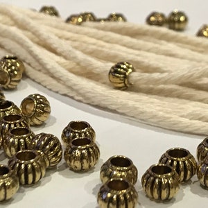 May include: A collection of small, round, metallic gold-toned beads with a vertical ribbed texture scattered alongside a length of thick, off-white twisted cotton cord. The beads feature a central hole and a shiny, reflective finish.