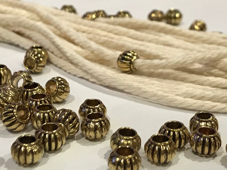 May include: A collection of small, round, metallic gold-toned beads with a vertical ribbed texture scattered alongside a length of thick, off-white twisted cotton cord. The beads feature a central hole and a shiny, reflective finish.