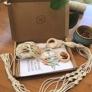 May include: A macrame plant hanger kit in a brown cardboard box with the text WILDE AT HOME. The kit includes off-white cotton cord, two light wood rings, and multi-colored wooden beads. An instruction sheet with a plant illustration is included.