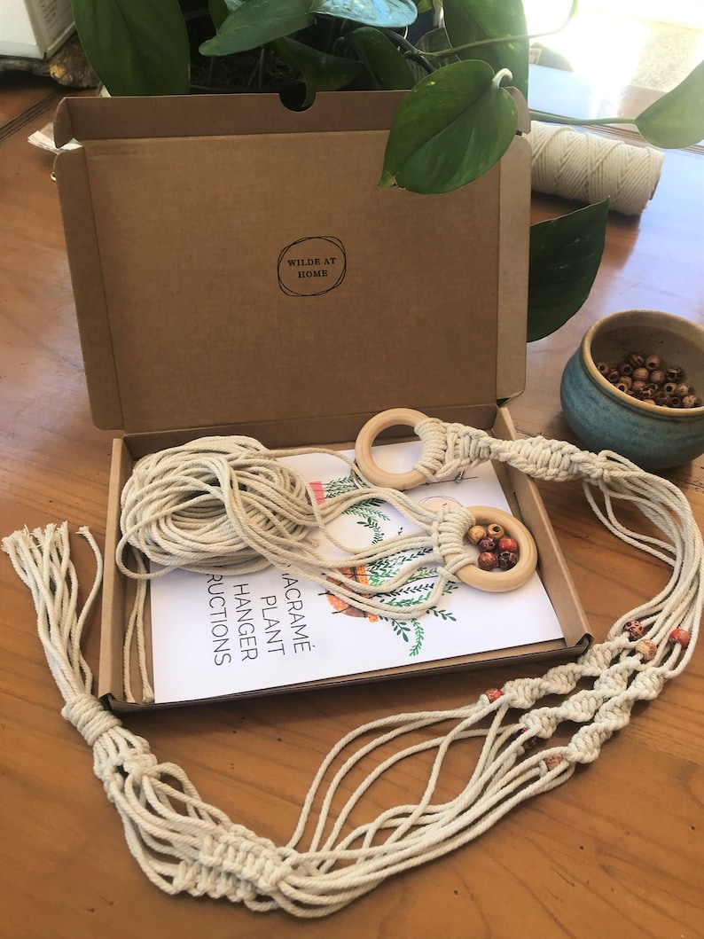 May include: A macrame plant hanger kit in a brown cardboard box with the text WILDE AT HOME. The kit includes off-white cotton cord, two light wood rings, and multi-colored wooden beads. An instruction sheet with a plant illustration is included.